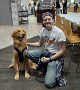 Man smiling and kneeling next to golden retriever in a cafeteria. Man is wearing a University of Virginia shirt.