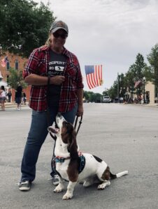 Christin Benoit standing with her basset on a street. There is an American flag behind her.