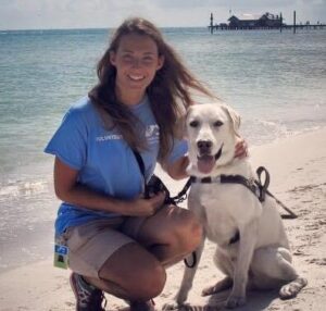 Kelsey Fernandez next to a yellow labrador guide dog on a beach.