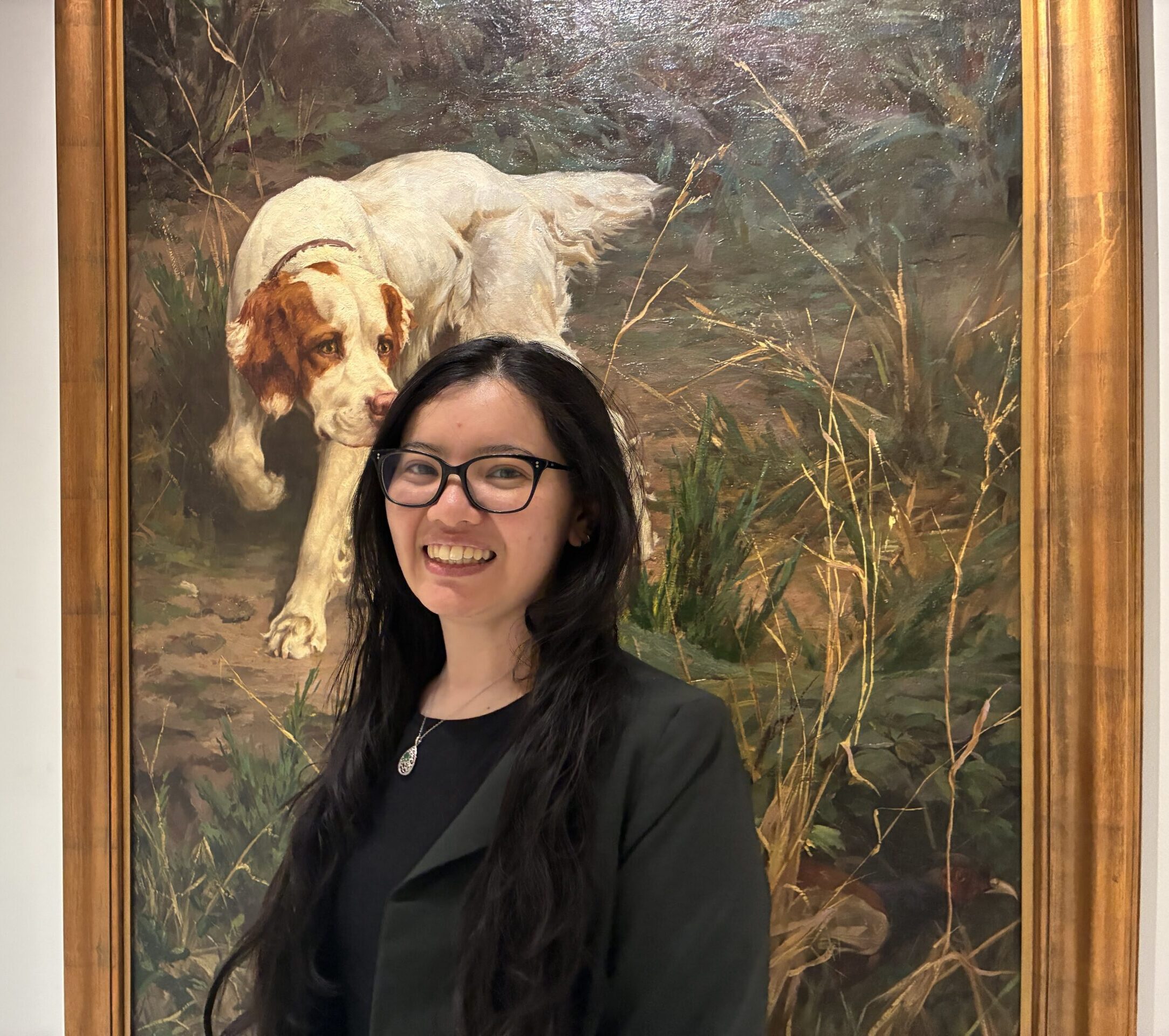 woman wearing glasses smiling at camera in front of painting of a dog
